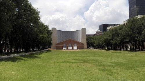 A grassy area with trees, featuring a modern building with a textured wall in the background under a cloudy sky.