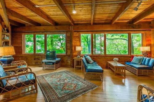 Cozy wooden living room with large windows, blue seating, a rug, and natural light from surrounding greenery.