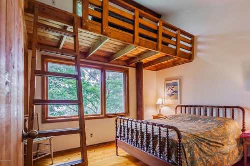 Cozy bedroom with a wooden loft, large window, and a bed with a decorative coverlet. Natural light fills the space.