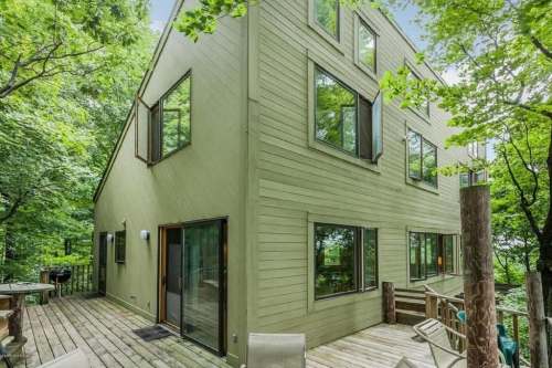 A modern green house surrounded by trees, featuring large windows and a wooden deck.