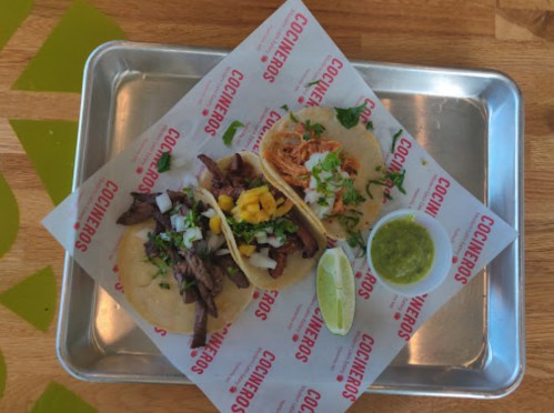 Two tacos on a tray: one with beef and the other with chicken, garnished with onions, cilantro, and lime, with green salsa.