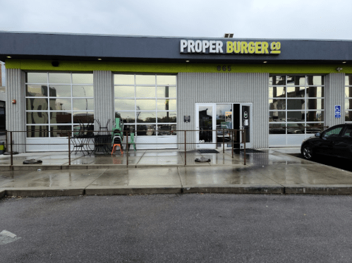 Exterior of Proper Burger Co. restaurant with large windows and a modern design, featuring a parking area in front.