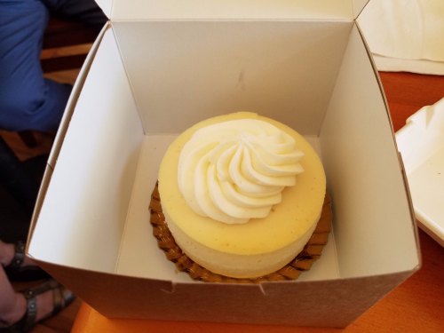 A creamy cheesecake with a swirl of whipped cream, placed in a brown takeout box on a wooden table.