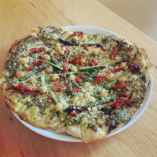A round pizza topped with pesto, sun-dried tomatoes, chickpeas, and herbs on a wooden table.