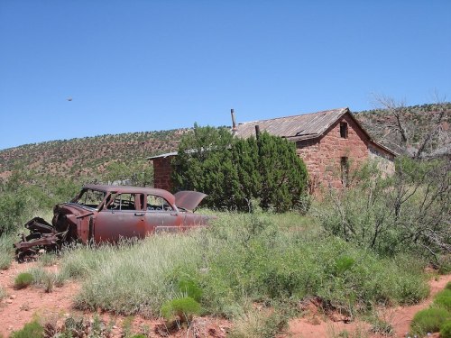 An old, rusted car sits in front of a weathered stone house, surrounded by overgrown grass and shrubs under a clear blue sky.