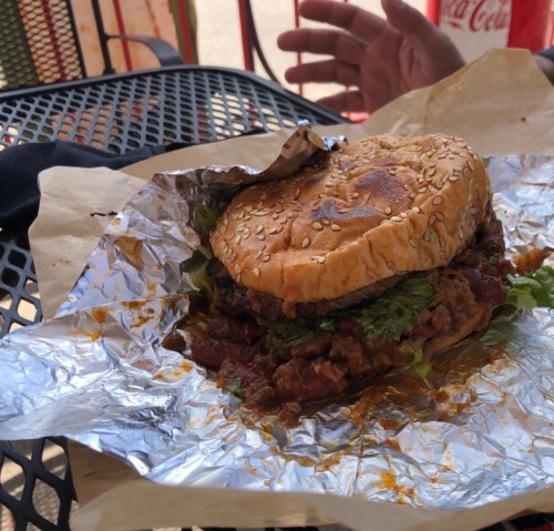 A sesame seed burger with lettuce and a thick patty, resting on foil, with a hand in the background.