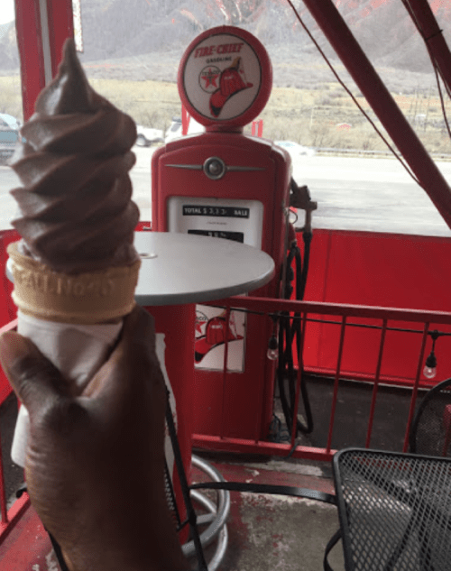 A hand holds a chocolate soft serve ice cream cone in front of a vintage gas pump and red outdoor seating.