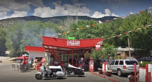 A retro-style diner with a red roof, surrounded by trees and mountains, featuring outdoor seating and parked motorcycles.