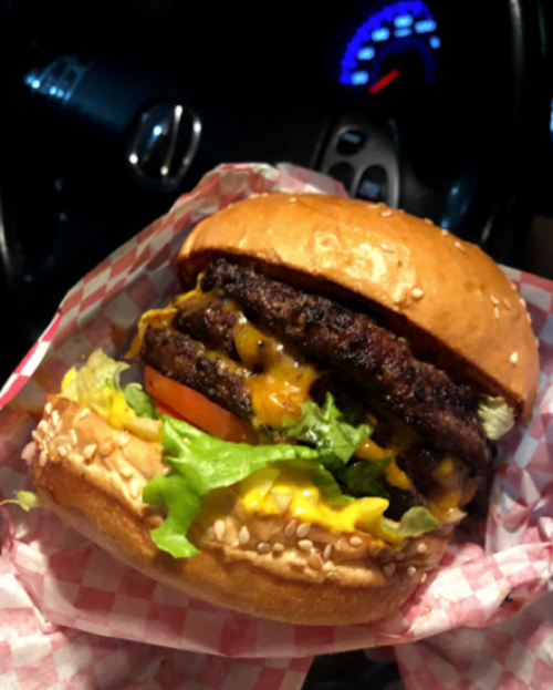 A close-up of a stacked burger with two patties, cheese, lettuce, tomato, and mustard in a sesame seed bun.