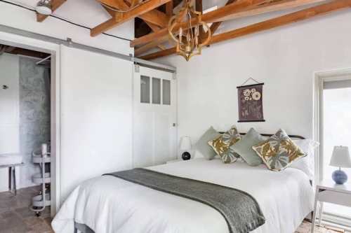 Cozy bedroom with a bed, decorative pillows, wooden beams, and a small lamp beside a window.