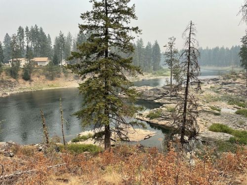A serene river scene surrounded by trees, with a smoky haze in the background and rocky banks.