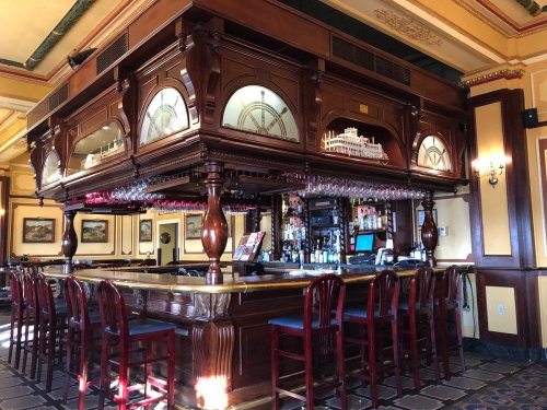A wooden bar with a decorative overhead structure, surrounded by high-backed chairs in a warmly lit pub.