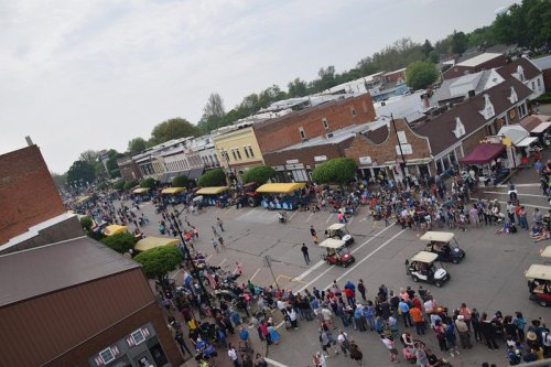 Aerial view of a busy street festival with crowds, golf carts, and vendor tents lining the road.