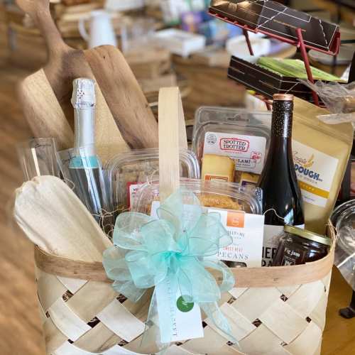 A woven basket filled with gourmet food items, a rolling pin, and a decorative bow, set on a wooden floor.