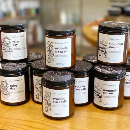 A collection of candle jars on a wooden surface, featuring labels like "Rainy Day" and "Avocado & Sea Salt."