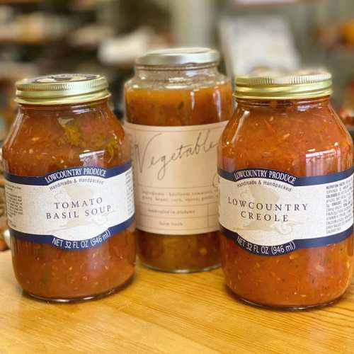 Three jars of soup: Tomato Basil, Vegetable, and Lowcountry Creole, displayed on a wooden surface in a cozy setting.