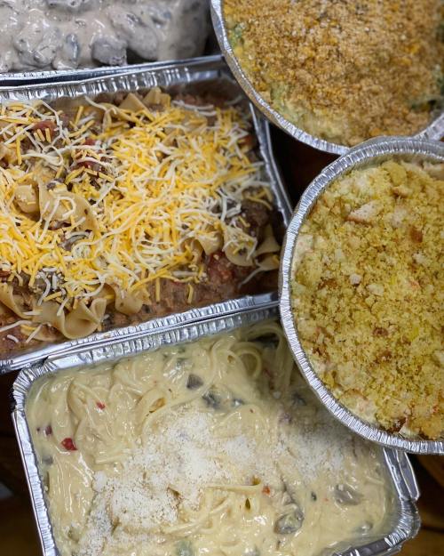 A variety of baked dishes in aluminum trays, including pasta, casseroles, and a creamy sauce dish, topped with cheese and breadcrumbs.
