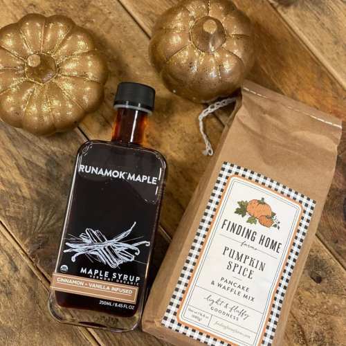 A bottle of maple syrup, a bag of pumpkin spice mix, and two gold decorative pumpkins on a wooden surface.