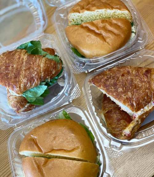 Four sandwiches in clear containers: a croissant sandwich with greens, and two round sandwiches, one cut in half.