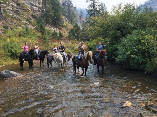 A group of people on horseback crossing a shallow stream surrounded by trees and rocky terrain.