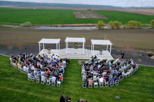 A wedding ceremony setup with guests seated in a heart shape around a white altar in a green landscape.
