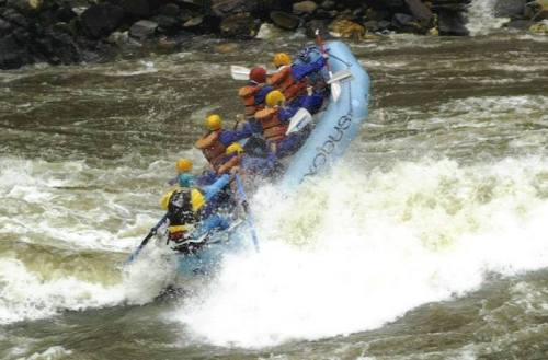 A group of people in life jackets navigating a white-water river in a blue raft, splashing through waves.