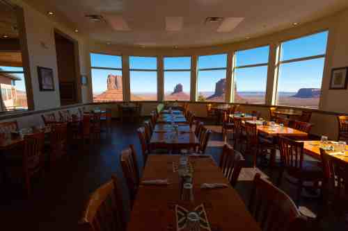 A restaurant interior with wooden tables and chairs, featuring large windows showcasing a stunning desert landscape.