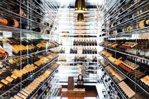 A modern wine cellar with glass walls, showcasing rows of wine bottles on sleek shelves.