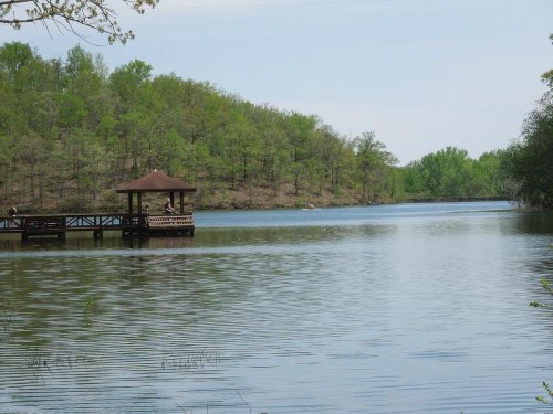 A serene lake surrounded by green trees, featuring a wooden gazebo on a dock.