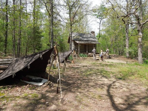 A rustic cabin in a wooded area, with two people near the entrance and tents set up nearby.