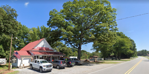 A small white building with a red roof, surrounded by trees and parked cars, along a rural road.