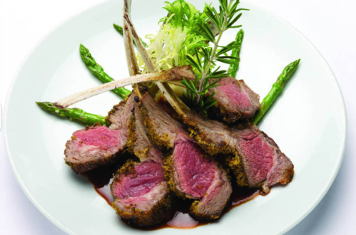 A plate of sliced lamb rack with herbs, asparagus, and a drizzle of sauce, elegantly presented.