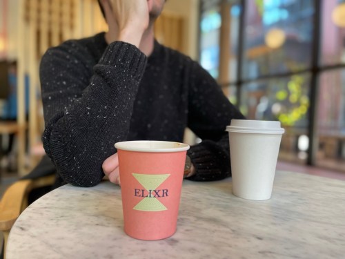 A person sits at a table with a pink coffee cup labeled "ELIXR" and a white cup, in a cozy café setting.