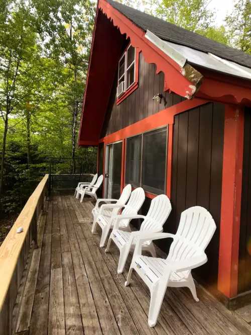 A cozy cabin with a wooden deck and white chairs, surrounded by lush green trees.