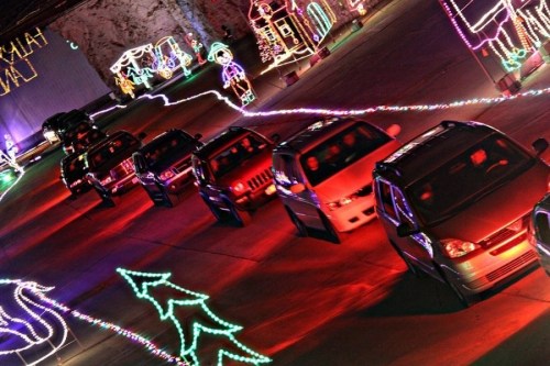 Cars driving through a festive display of colorful holiday lights in a dark setting.