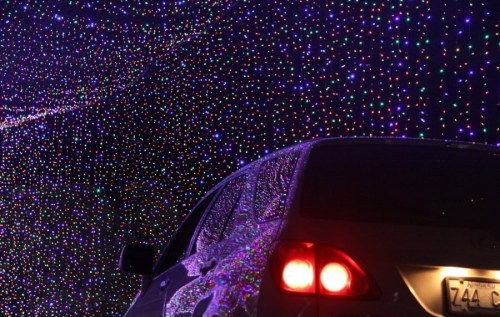 A car parked under a colorful display of twinkling holiday lights.
