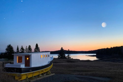 A boat-shaped structure sits by a calm lake at sunset, with a bright moon and stars in the clear sky.