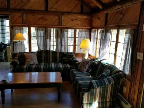 Cozy living room with wooden walls, striped sofas, a coffee table, and large windows with sheer curtains.