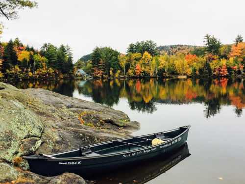 A calm lake surrounded by autumn foliage, with a black canoe resting on a rocky shore.