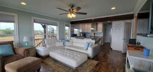 Bright living room with a white sectional sofa, ceiling fan, and open kitchen, featuring large windows and wooden floors.