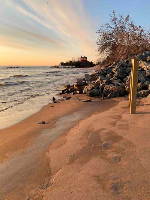 A serene beach at sunset, with gentle waves, rocks, and a distant house surrounded by trees.