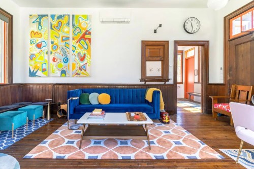 Bright living room featuring a blue sofa, colorful artwork, wooden accents, and a patterned rug.