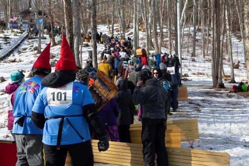 A crowd of people in winter attire, some wearing red hats, walking along a snowy path in a wooded area.