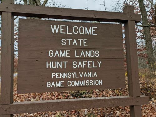 Wooden sign reading "Welcome State Game Lands Hunt Safely Pennsylvania Game Commission" with autumn foliage in the background.