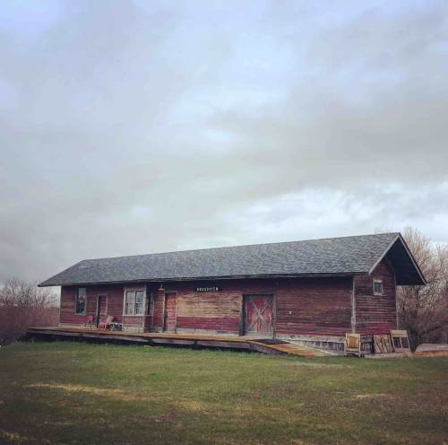 A rustic wooden house with a gray roof, set on a grassy area under a cloudy sky.