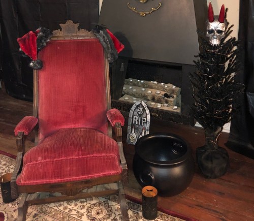 A spooky scene featuring a red chair, a black tree with a skull, a cauldron, and a tombstone labeled "RIP."