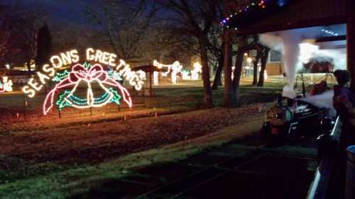 A festive display with "Seasons Greetings" lights and a small train emitting steam, set in a park at night.