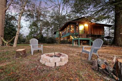 A cozy cabin surrounded by trees, featuring a fire pit and outdoor seating in a serene setting at dusk.