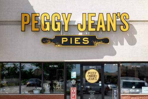 Sign for "Peggy Jean's Pies" featuring a rolling pin graphic, with storefront windows displaying "Open" and "Welcome."