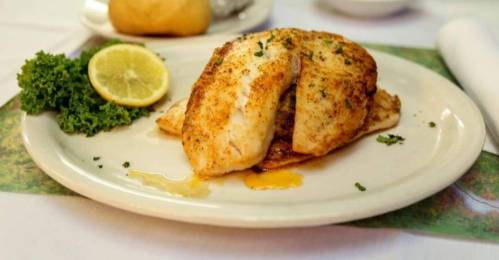 A plate of golden-brown fish fillet garnished with herbs, served with a lemon wedge and a side of kale.